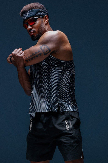 A man in athletic wear posing in a studio setting. He is wearing a black and gray patterned sleeveless tank top, black athletic shorts, a black headband, and blue running shoes against a dark blue-gray background.