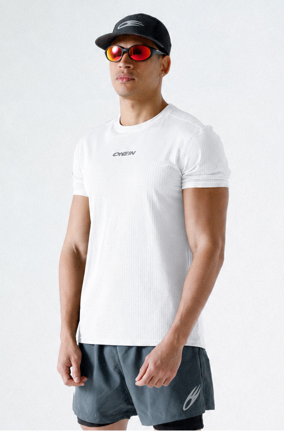 A man modeling athletic wear, featuring a white short-sleeve t-shirt with a small black logo on the chest, dark gray athletic shorts, a black cap, and black sneakers, set against a plain light gray studio background.