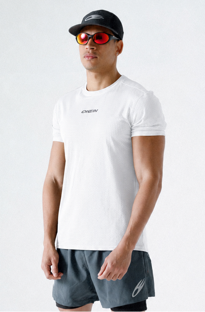 A man modeling athletic wear, featuring a white short-sleeve t-shirt with a small black logo on the chest, dark gray athletic shorts, a black cap, and black sneakers, set against a plain light gray studio background.
